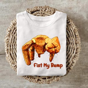 May include: White t-shirt featuring a graphic of a fist bump in shades of brown and orange. The text "Fist My Bump" is printed in a reddish-brown color below the graphic. The shirt is displayed on a woven basket.