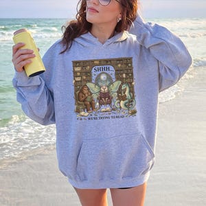 May include: A light gray hooded sweatshirt featuring a graphic print of a library scene with a moth, a sasquatch, and a dragon reading books. The text reads SHHH... WE'RE TRYING TO READ. The sweatshirt has long sleeves, a front kangaroo pocket, and a hood.