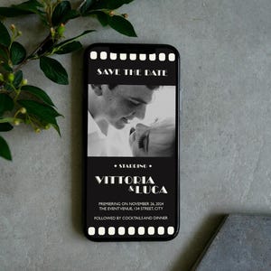 May include: Black and white photo of a couple on a smartphone screen with a film strip border. The text "Save the Date" is at the top of the screen. The text "Starring Vittoria & Luca" is below the photo. The text "Premiering on November 26, 2024 The Event Venue, 134 Street, City" is below the names. The text "Followed by Cocktails and Dinner" is at the bottom of the screen.