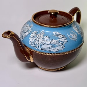 Puede incluir: Una tetera de cerámica marrón y azul con tapa y asa. La tetera presenta una banda azul claro con figuras clásicas blancas y diseños de paisajes. Los bordes y el pico están adornados con detalles dorados.
