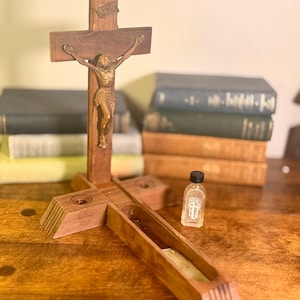 Puede incluir: Un crucifijo de madera con una figura de Jesús de bronce, colocado sobre una base de madera con un canal. Una pequeña botella con un emblema de cruz está al lado. El fondo presenta una pila de libros.