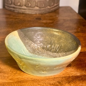 Puede incluir: Un cuenco de cerámica hecho a mano con un degradado de color único. El cuenco pasa de un verde pálido a un gris-marrón oscuro, con un borde amarillo. Las palabras "Love you" están inscritas en el lateral. El cuenco está sobre una superficie de madera.
