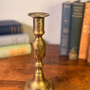 May include: A vintage brass candlestick holder with a hexagonal base and a detailed, ornate design. The candlestick is a warm gold color and stands upright on a wooden surface. Books are in the background.
