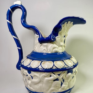 May include: A blue and white ceramic pitcher with a decorative handle and spout. The pitcher features a raised design of figures and foliage.