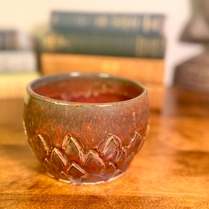 May include: A handmade ceramic bowl with a textured, floral-like pattern around the base. The bowl has a reddish-brown glaze with darker accents, and a smooth rim. The bowl is sitting on a wooden surface.