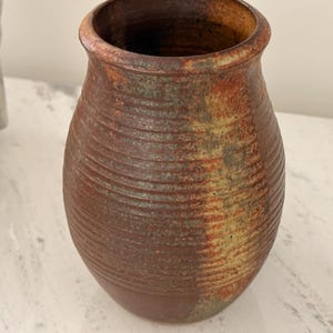 May include: A cylindrical ceramic vase with a rounded body and a flared rim. The exterior features a textured surface with horizontal ridges and a variegated glaze in shades of deep brown, rust orange, and muted green, creating a mottled appearance.