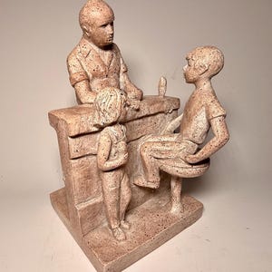 Op de afbeelding: Een keramische sculptuur van drie figuren, een man en twee kinderen, die aan een toonbank staan. De man draagt een shirt met korte mouwen en de kinderen dragen casual kleding. De sculptuur is geschilderd in tinten bruin en wit.