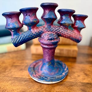 Puede incluir: Un candelabro de cerámica hecho a mano con cinco candeleros. La pieza presenta una vibrante paleta de colores azul, morado y dorado. La base es redonda y los brazos se extienden hacia afuera, creando un diseño único. Un artículo decorativo para el hogar.