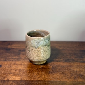 Puede incluir: Una taza de cerámica hecha a mano con una superficie texturizada. La taza presenta una combinación de colores beige y verde, una base redondeada y un borde ligeramente ensanchado. Para bebidas.