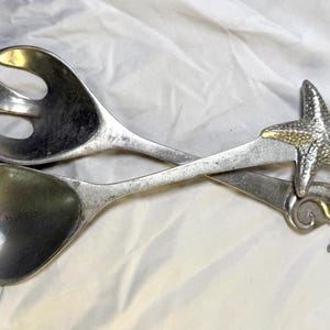 Könnte beinhalten: Silberfarbene Salatbesteck mit einem Seestern- und Seepferdchen-Design. Der Servierlöffel und die Gabel sind gekreuzt und liegen auf einer weißen Oberfläche. Der Seestern ist strukturiert und fügt ein maritimes Thema hinzu.