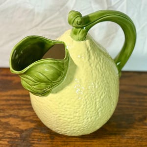 Puede incluir: Jarra de cerámica con forma de limón, con cuerpo amarillo texturizado y asa y boquilla verdes. La boquilla está diseñada para parecerse a una hoja. Este artículo es un objeto decorativo.