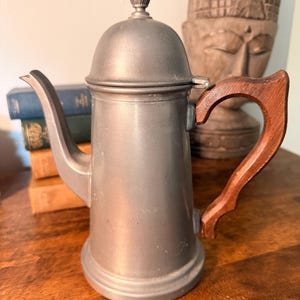 May include: A vintage pewter coffee pot with a wooden handle. The pot has a domed lid with a pineapple finial. The handle is a dark brown color. The pot is sitting on a wooden surface.