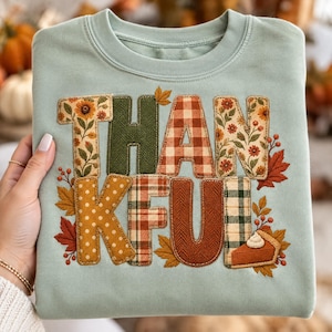May include: A light sage green crewneck sweatshirt featuring the word THANKFUL in large, appliqued letters. Each letter displays a different autumn-themed pattern, including floral prints, green herringbone, orange gingham, and brown textures.