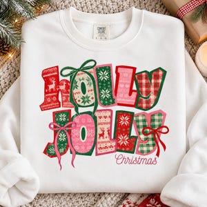 May include: A white crewneck sweatshirt featuring the text Holly Jolly Christmas in large, colorful block letters. The letters are decorated with red, green, and pink patterns including reindeer, snowflakes, and plaid, accented by green and pink bows.