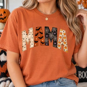 May include: An orange short-sleeve t-shirt featuring the word MAMA printed across the chest. Each letter has a distinct Halloween pattern: ghosts, bats, spiderwebs with spiders, and candy corn. The letters are outlined with a visible stitched border.