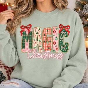 May include: A sage green crewneck sweatshirt featuring the word MAGIC in large, patterned block letters decorated with red bows, plaid, candy canes, and ornaments. Below, the word Christmas is written in a cursive, light pink font.