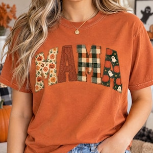 May include: A burnt orange short-sleeve t-shirt featuring the word MAMA in large block letters. Each letter has a different pattern: floral with pumpkins, solid brown cable knit, green and black buffalo plaid, and dark green with ghosts.