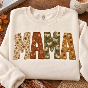 Könnte beinhalten: Ein cremefarbenes Sweatshirt mit Rundhalsausschnitt und dem Wort MAMA in großen, aufgenähten Stoffbuchstaben. Jeder Buchstabe zeigt ein florales Muster in Senfgelb, Rostorange, Salbeigrün und Weiß mit deutlich sichtbaren Nähten.