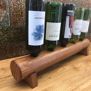 May include: A wooden wine rack holds five wine bottles. The rack is made of light brown wood and has a cylindrical design. The bottles are various colors, with labels visible. The rack is on a wooden surface.
