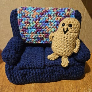 May include: A crocheted couch with a blue fabric and a colorful blanket. A crocheted brown figure with a smiling face sits on the couch.