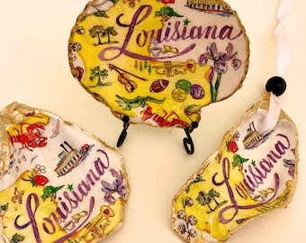May include: Hand-painted oyster shells with a Louisiana theme. The shells feature yellow backgrounds with purple lettering that spells "Louisiana". The shells are decorated with images of a fleur-de-lis, a crawfish, and a steamboat.