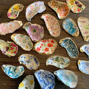 May include: A collection of 18 decorative oyster shells, each painted with a different floral pattern. The shells are arranged in a circle on a wooden surface.
