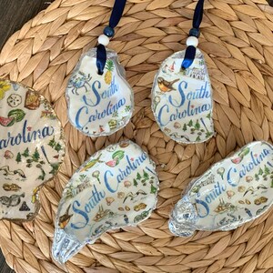 May include: Four oyster shells decorated with decoupage illustrations of South Carolina landmarks, including a crab, pineapple, and lighthouse. The shells feature the text South Carolina in blue script, with two shells hanging from dark blue ribbons.