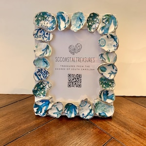 May include: A white picture frame decorated with blue and white oyster shells. The frame has a white mat and a white background with the text "SCCOASTAL TREASURES TREASURES FROM THE SHORES OF SOUTH CAROLINA" and a QR code.