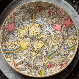 May include: A decorative ceramic plate with a floral design. The plate features a yellow flower in the center, surrounded by pink tulips, blue and yellow accents, and green foliage. The rim is trimmed with gold.