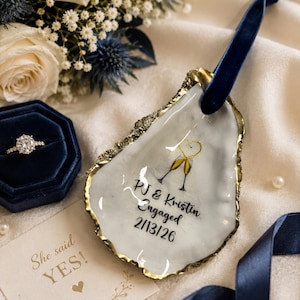 May include: An oyster shell ring dish with gold trim, featuring the text "PJ & Kristin Engaged 2/13/26" and two champagne glasses. A blue velvet ribbon is attached. A diamond ring in a blue box and a card that says "She said YES!" are also present.