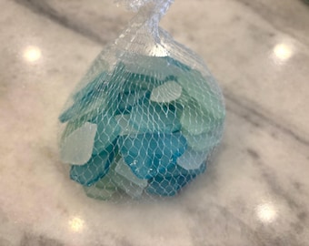 May include: A mesh bag filled with sea glass in shades of blue and white. The sea glass pieces have smooth, rounded edges and vary in size. The bag is tied at the top with a white string, and the background is a marble surface.