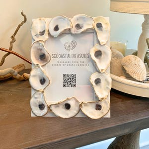 May include: A white picture frame decorated with oyster shells. The frame has the text "SC COASTAL TREASURES" and a QR code. The oyster shells are white with a dark brown interior.