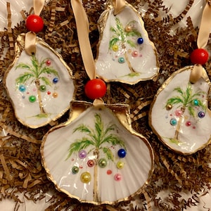 May include: Hand-painted oyster shell ornaments with a gold rim, featuring a palm tree design and colorful bead accents. Each ornament has a beige ribbon and a red bead for hanging. The shells are displayed on a white plate.