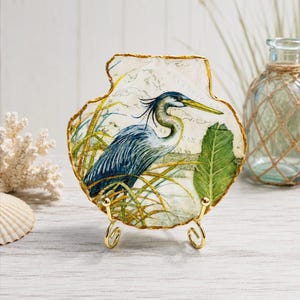 May include: A decorative scallop shell featuring a printed illustration of a blue heron standing among tall grasses and a large green leaf. The shell has a gold-painted scalloped edge and is displayed on a small gold-toned metal wire stand.