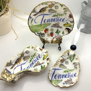 May include: Three decorative oyster shells with a Tennessee theme. Each shell is painted with a map of Tennessee, a bird, and other Tennessee-related images. The shells have gold-painted edges and are on a white surface.