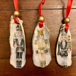 May include: Three Christmas ornaments featuring nutcracker soldier designs. Each ornament is painted on a white oyster shell, with a red ribbon and gold bead for hanging. The nutcrackers are dressed in black, white, and gold uniforms.