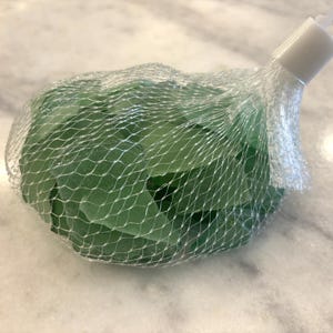 May include: A mesh bag filled with sea glass pieces. The sea glass is a translucent green color. The bag has a white plastic closure and a white string for hanging. The background is a light-colored, marble-like surface.
