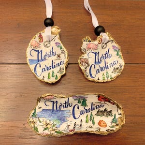 May include: Three oyster shell ornaments with gold accents and a white background. Each ornament features a hand-painted illustration of North Carolina with various landmarks and the text "North Carolina".