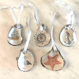 May include: Five oyster shell ornaments with different nautical designs. The ornaments are painted with a sailboat, a wheel, an anchor, a starfish, and a seagull. Each ornament has a white ribbon attached.