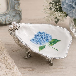 May include: A white oyster shell dish featuring a hand-painted blue hydrangea flower in the center. The shell has a metallic silver rim and three small silver legs. Three white pearls are attached to the top edge of the shell.