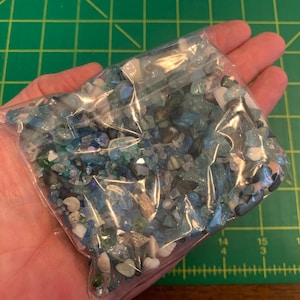May include: A clear plastic bag filled with small, irregularly shaped blue, white, and clear glass pieces. The bag is held in a person's hand against a green cutting mat.