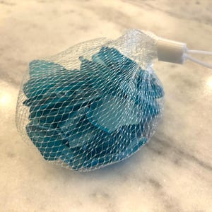 May include: A mesh bag filled with small, irregular-shaped, teal-colored glass pieces. The bag has a white plastic closure with a loop for hanging. The glass pieces are visible through the netting.