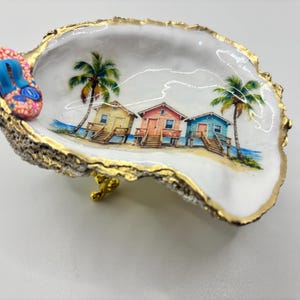May include: A decorative oyster shell dish with a gold-painted rim and a small, colorful flip-flop charm attached to the edge. The interior features a painted scene of three beach houses in yellow, pink, and blue, flanked by two palm trees.