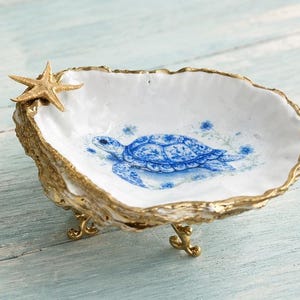 May include: A decorative, white ceramic dish with a blue turtle design and gold trim. A gold starfish adorns the edge. The dish is supported by three gold feet, creating a coastal or nautical theme.