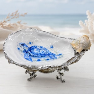 May include: A decorative oyster shell dish with a blue sea turtle design. The shell is set on a silver-colored metal stand and features a small seashell on the side. The background shows a beach scene with the ocean and coral.