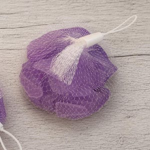 May include: A collection of small, smooth, translucent purple glass pieces contained within a white mesh netting bag. The bag is secured at the top with a white plastic fastener and features a thin white cord loop for hanging or attachment.