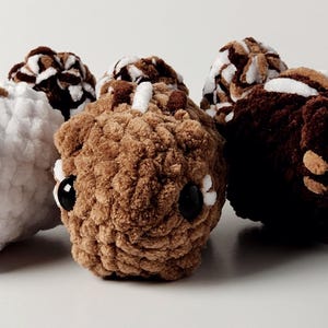 May include: Three crocheted plush toys in the shape of animals. The first is white with brown accents, the second is brown with white accents, and the third is brown. All three have large black eyes.