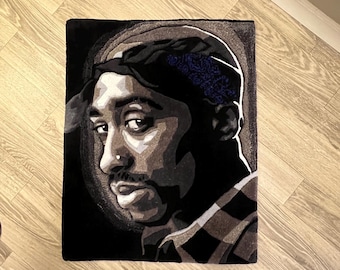 May include: A black and white rug with a portrait of a man with a bandana. The man's face is the focal point of the rug, and his eyes are looking directly at the viewer. The rug is made of a soft, plush material.