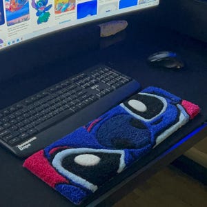 May include: A blue, red, and white Stitch-themed rug, featuring the character's face. The rug is rectangular and sits on a black desk next to a keyboard and mouse. The computer screen displays various Stitch images.