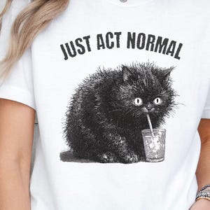 May include: White t-shirt with the text "JUST ACT NORMAL" above a black cat illustration. The cat is drinking from a glass with a straw. The shirt is a casual, everyday wear item.